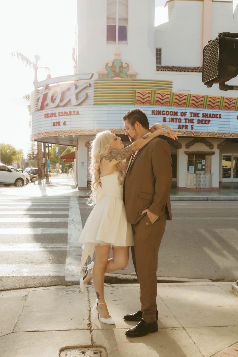 Jacinda in a white dress kicks up a heel while embracing Matthew in a brown suit on a downtown Bakersfield sidewalk.