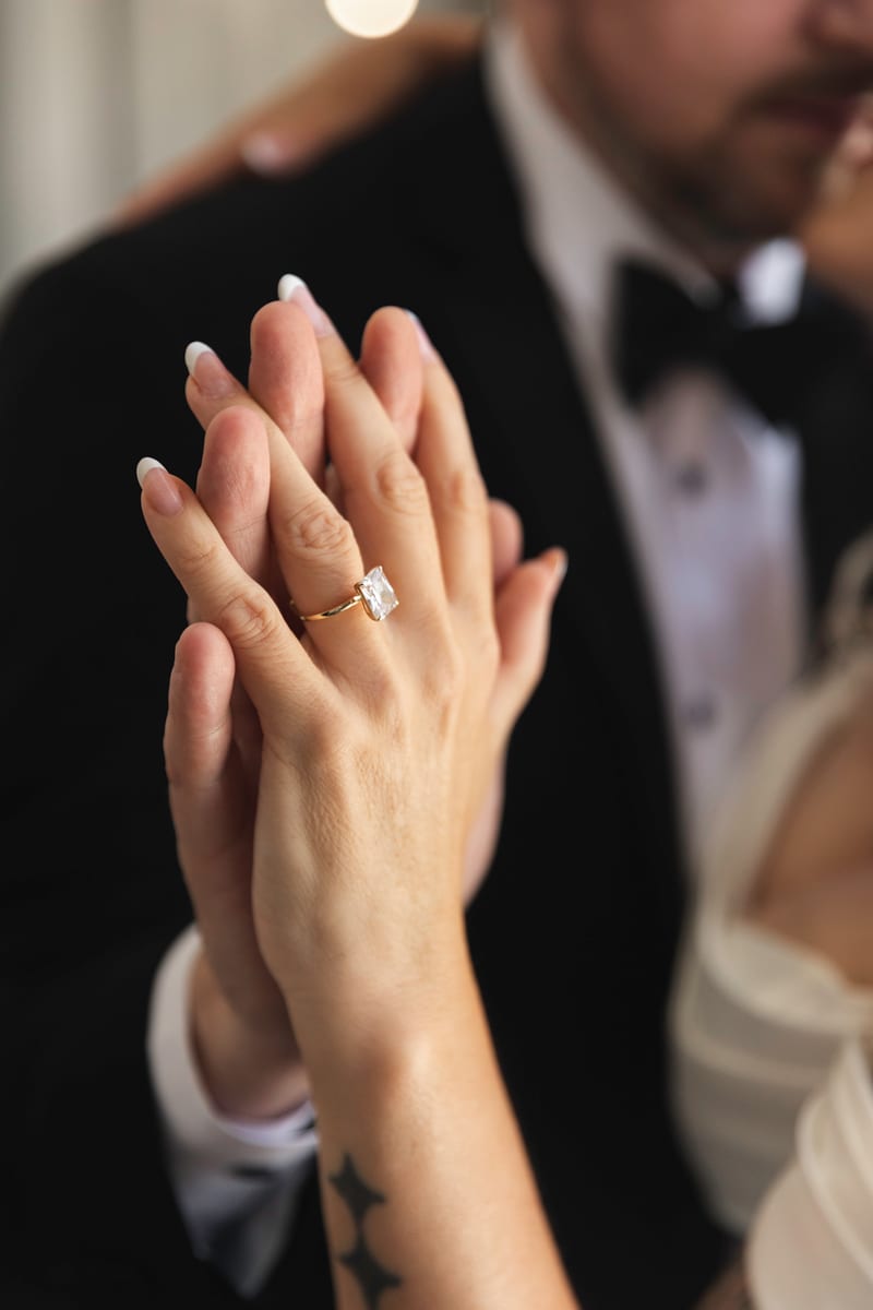 Clasped hands with an emerald-cut diamond ring, French manicure, and star tattoo, tuxedo blurred behind